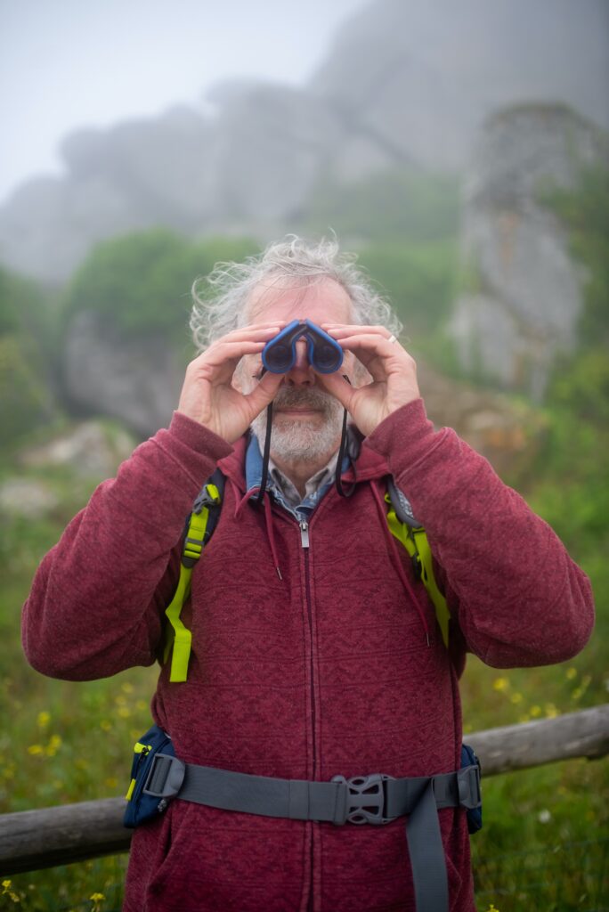 Gray-haired man looking at the camera with binoculars with mountain in background